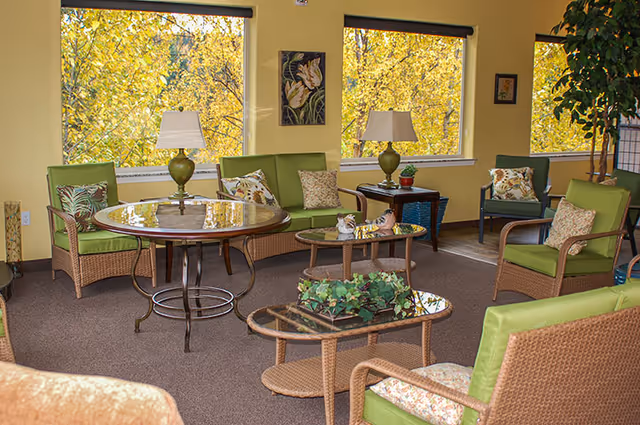 A cozy living room area with wicker furniture featuring green cushions and floral pillows. There are two round glass-top tables with decorative items and two table lamps. Large windows show a view of trees with yellow leaves outside, and the walls are painted yellow with framed artwork.