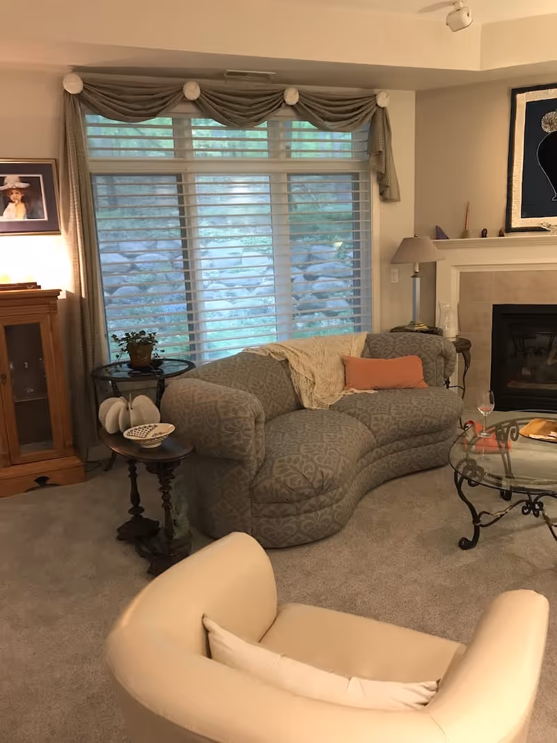 Cozy living room with a curved patterned sofa, armchair, glass coffee table, fireplace and large window with blinds.