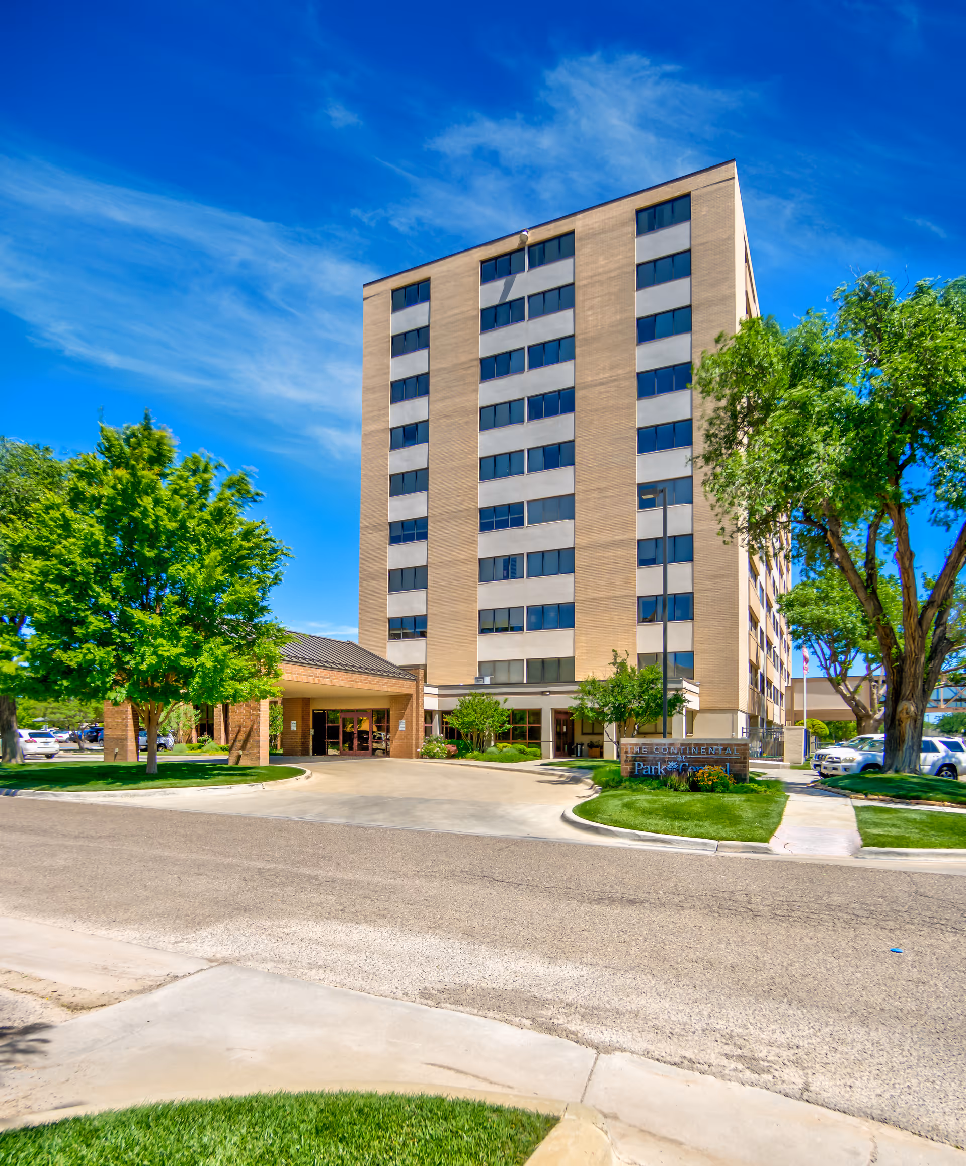 Front exterior of a multi-story senior living building with a covered entrance, driveway, lawn and trees under a blue sky.