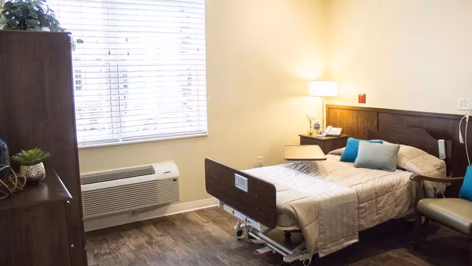 A well-lit bedroom in an assisted living facility featuring a hospital-style bed with beige bedding and blue accent pillows. The room includes a wooden nightstand with a lamp, a phone, and a small plant. There is a window with white blinds, a wooden dresser with decorative items, and a beige armchair with a blue pillow. The floor is wood-style, and the walls are painted light yellow.