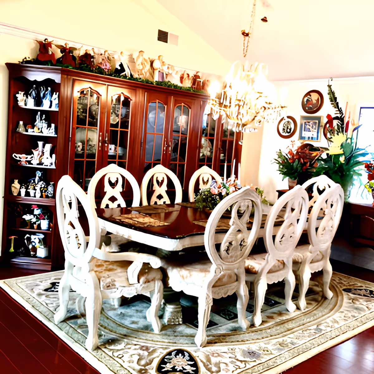 A formal dining room with a large wooden dining table surrounded by eight ornate white chairs. A chandelier hangs above the table. Behind the table is a large wooden cabinet with glass doors displaying various decorative items. The room is decorated with plants, framed pictures on the wall, and a patterned area rug under the table.