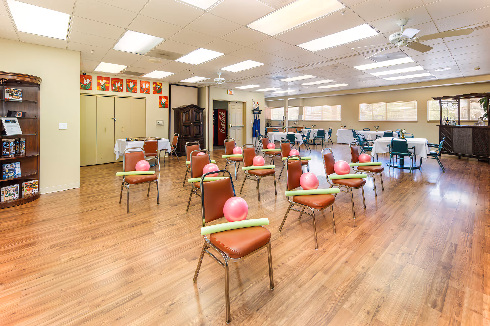 A spacious activity room with wooden flooring and multiple chairs arranged in rows, each chair holding a pink exercise ball and a green foam noodle. The room has several tables with chairs around them, a wooden cabinet, a vending machine, and large windows letting in natural light. The ceiling has fluorescent lights and a ceiling fan.