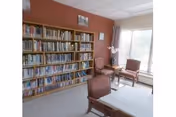 Interior view of a room with a large bookshelf filled with books along one wall, several chairs arranged near a window with natural light coming in, and a table in the foreground. The walls are painted a warm reddish color and there are framed pictures hanging above the bookshelf.