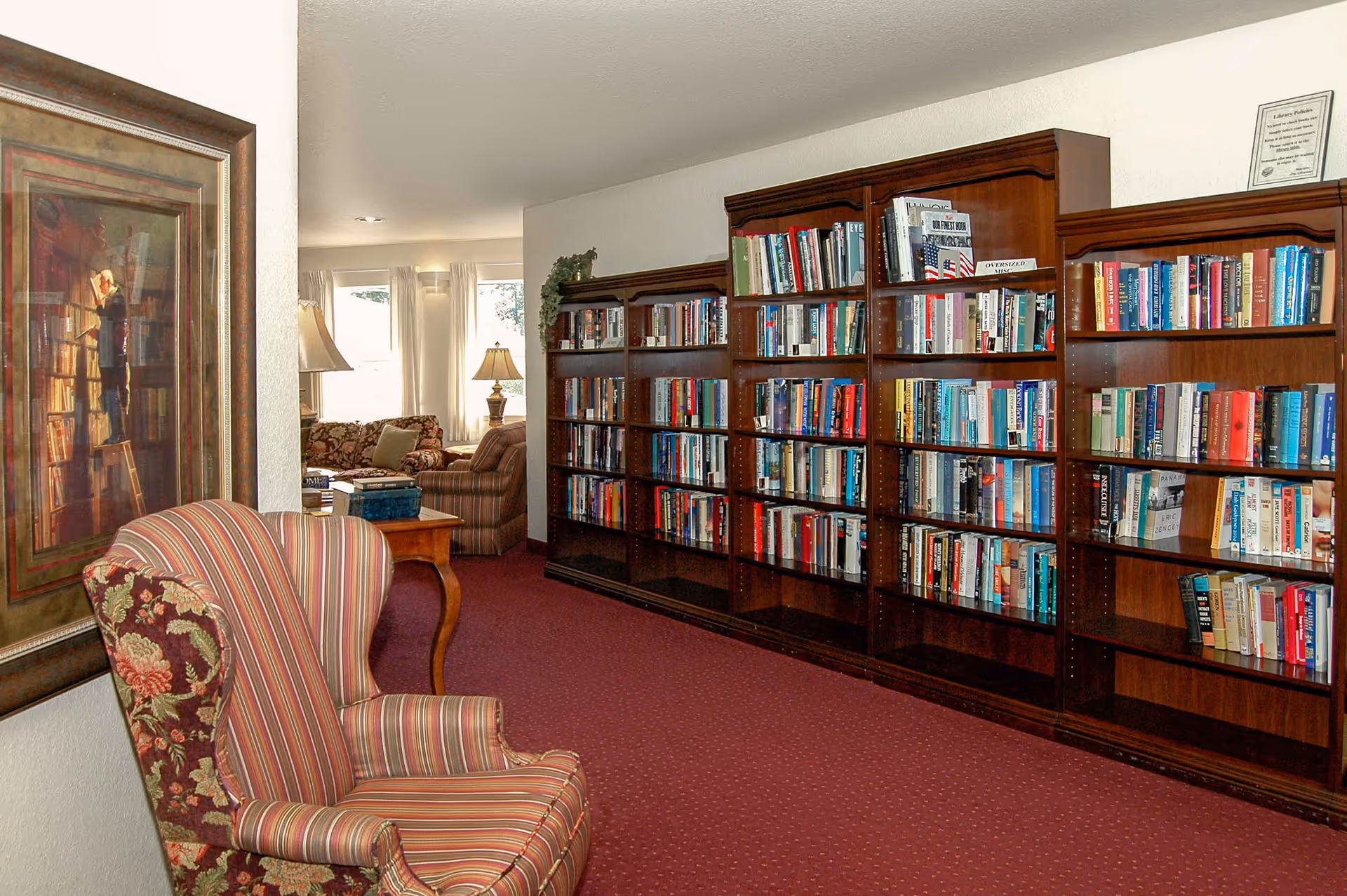 Interior view of a cozy library or reading area in a senior living facility. There are large wooden bookshelves filled with books along the right wall. A striped upholstered armchair with floral patterned sides is positioned near a framed painting on the left wall. In the background, there is a sitting area with a floral sofa, a striped armchair, a wooden side table with books, and two table lamps near windows with white curtains.