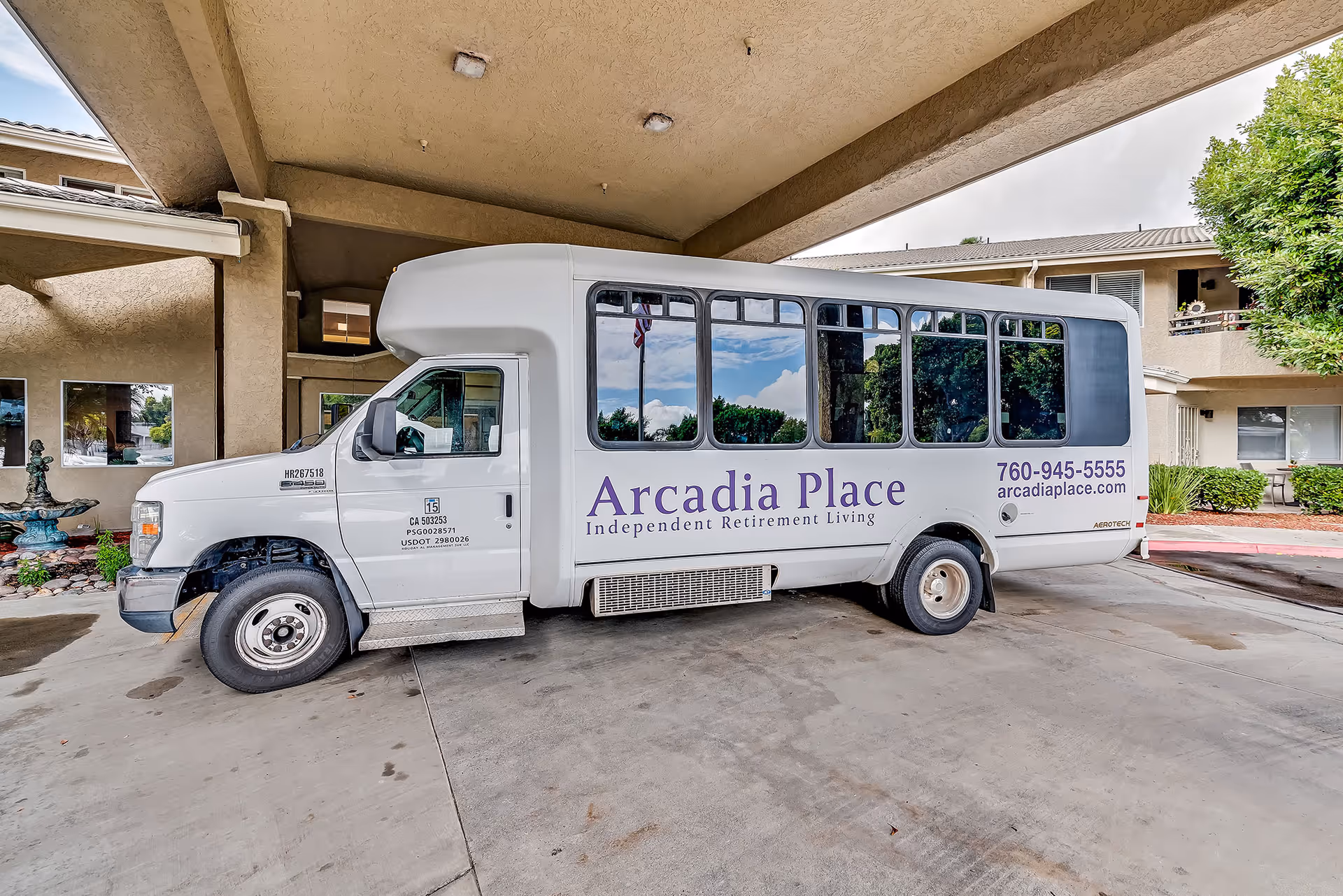 A white shuttle bus parked under a covered driveway at a senior living facility. The bus has large windows and displays the text 'Arcadia Place Independent Retirement Living' along with a phone number and website. The building and some greenery are visible in the background.