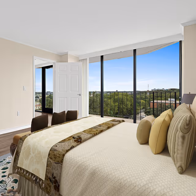 A bright bedroom with a large bed adorned with beige and patterned pillows and a decorative runner. The room features a large window wall with sliding glass doors that open to a balcony overlooking a green landscape and distant buildings under a clear blue sky. There is an open white door leading to another room with dark flooring.