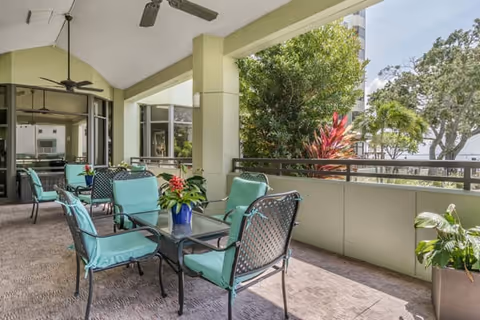 Covered outdoor patio area with several metal tables and chairs featuring turquoise cushions. The patio overlooks lush greenery and trees, with a ceiling fan mounted above for ventilation.