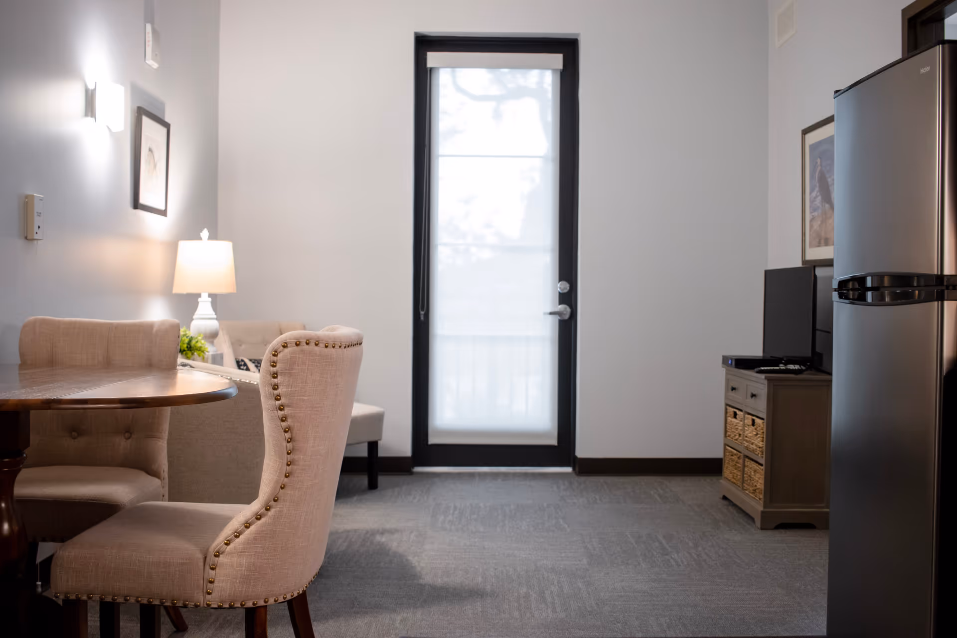 Interior view of a room with a round wooden table and upholstered chairs on the left, a lamp on a side table, a door with a window covered by a translucent blind in the center, and a refrigerator and a small cabinet with a TV on the right.