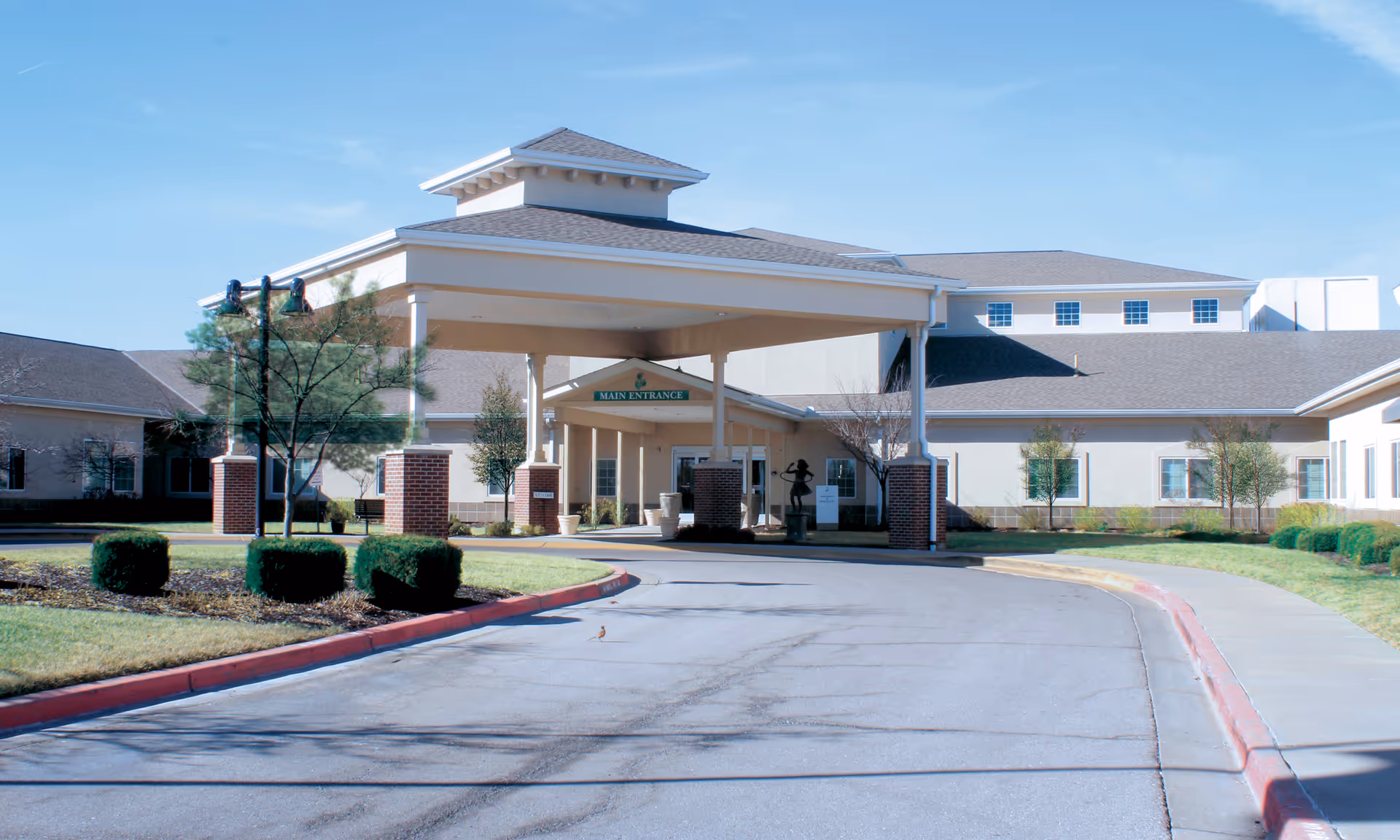 Exterior view of Pegasus Landing of Overland Park showing the main entrance with a covered driveway, brick pillars, and surrounding landscaping including bushes and trees under a clear blue sky.