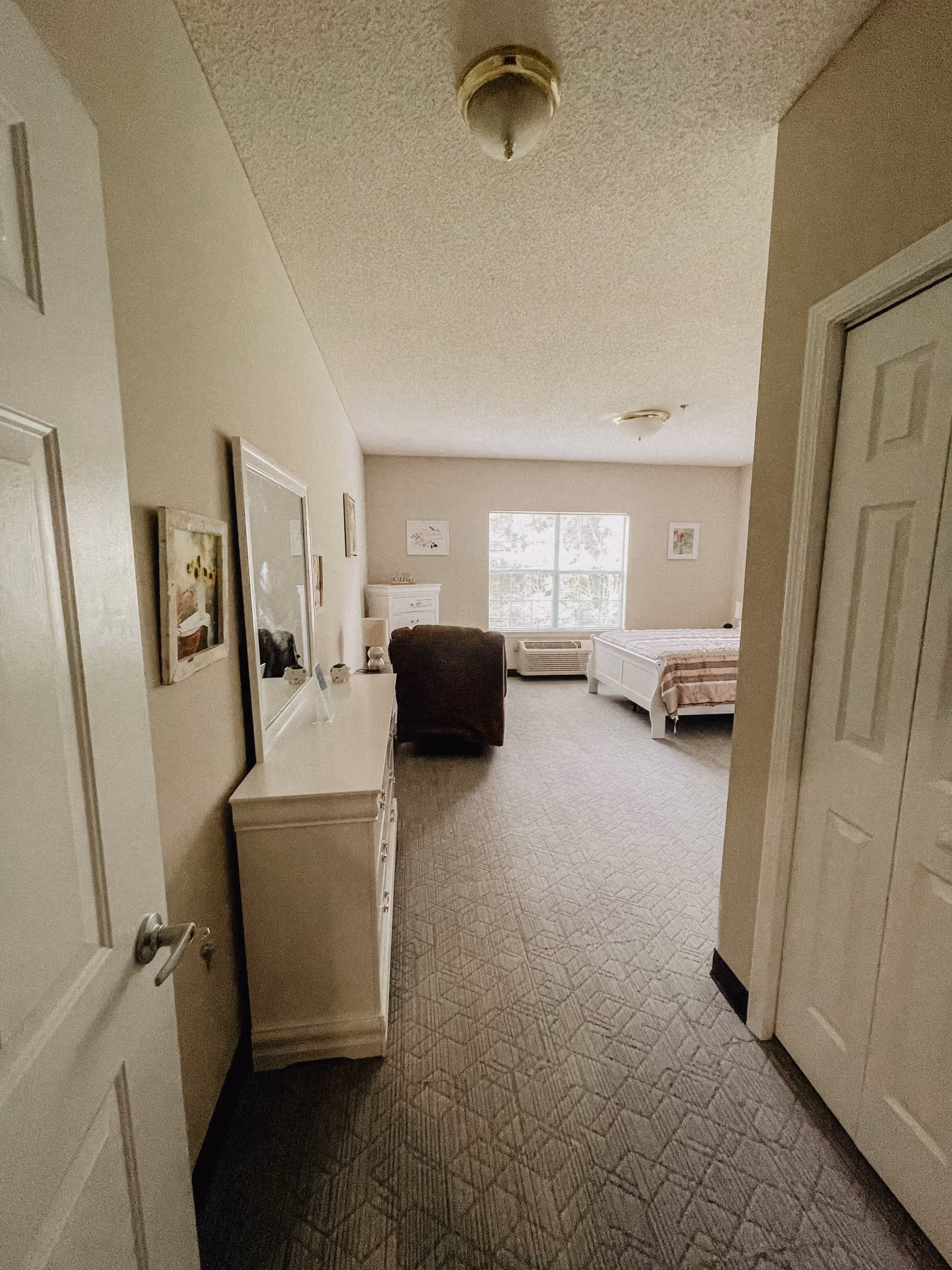 View down a hallway into a bedroom with a large window letting in natural light. The room contains a bed with a striped blanket, a brown armchair, a white dresser with a mirror, and framed pictures on the walls. The carpet has a geometric pattern and the walls are painted beige.