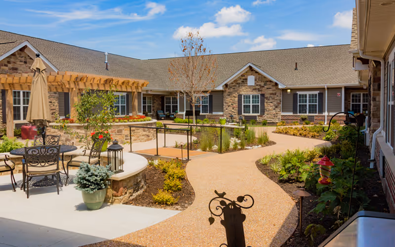 Outdoor courtyard area at a senior living facility with a winding paved pathway, garden beds, patio tables with chairs and umbrellas, and a pergola. The building surrounding the courtyard has stone and siding exterior with multiple windows.