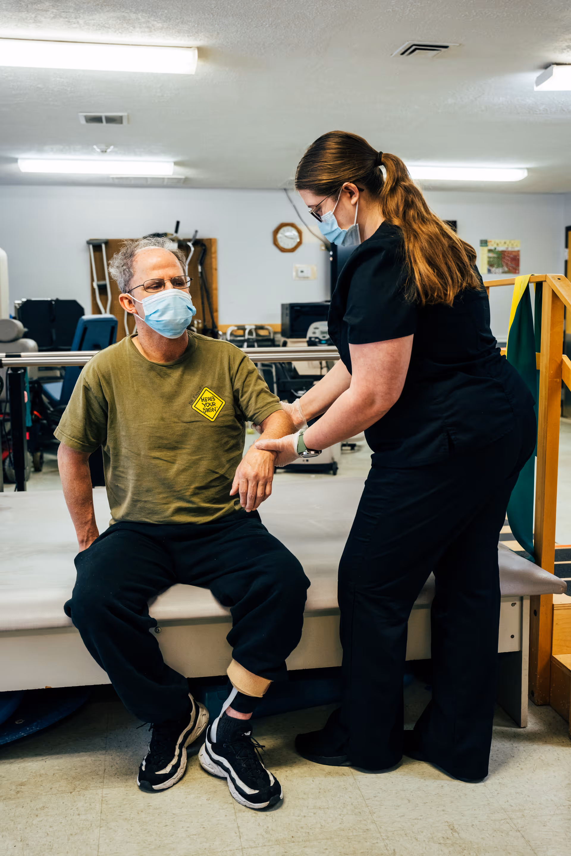 A man wearing a green t-shirt and black pants sits on a padded bench in a rehabilitation or physical therapy room while a female healthcare worker in black scrubs assists him by holding his arm. Both are wearing face masks. The room contains exercise equipment and has fluorescent lighting.