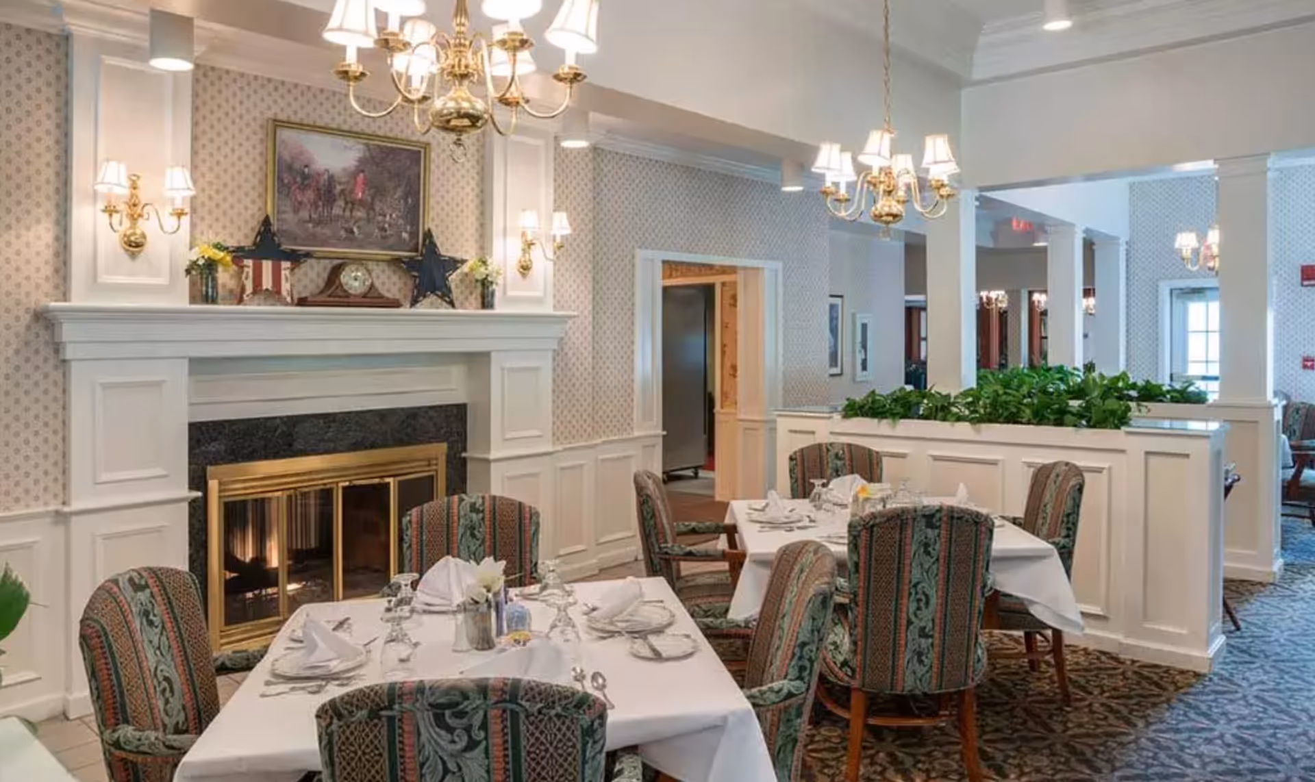 Elegant dining room with set tables and upholstered chairs in front of a white mantel fireplace and brass chandeliers.