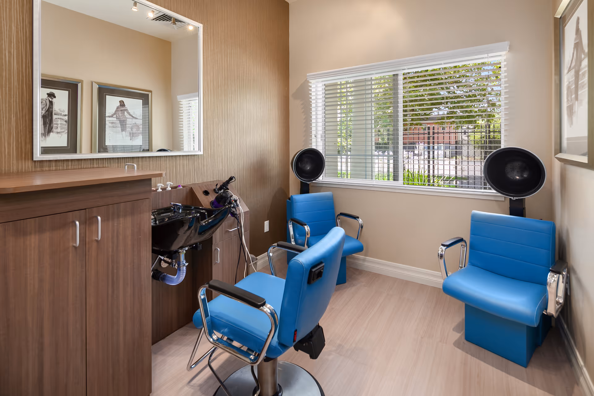 A small hair salon area with two blue salon chairs and two black hair dryers mounted on stands near a window with white blinds. There is a wooden cabinet with a black sink and a large mirror above it. The room has beige walls and light wood flooring.