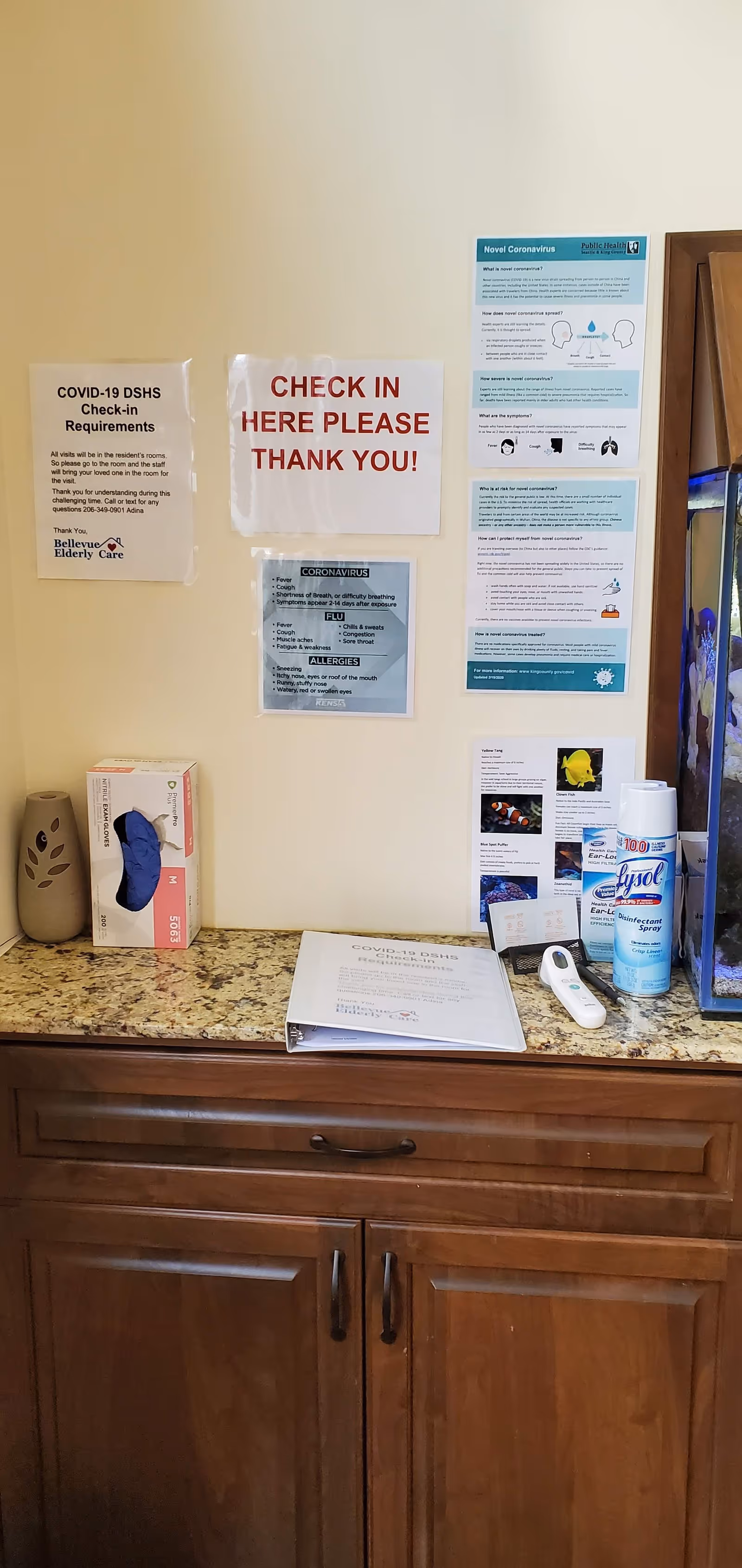 A check-in station at Bellevue Elderly Care with COVID-19 related signs on the wall including check-in instructions and coronavirus information. On the countertop, there are boxes of gloves, a thermometer, disinfectant wipes, and a binder labeled 'COVID-19 Guide'.