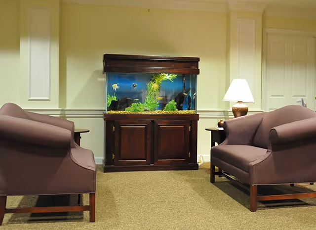 A cozy interior room with two purple upholstered armchairs facing each other, a wooden cabinet with a large fish tank filled with plants and fish in the center, and a table lamp on a side table to the right.