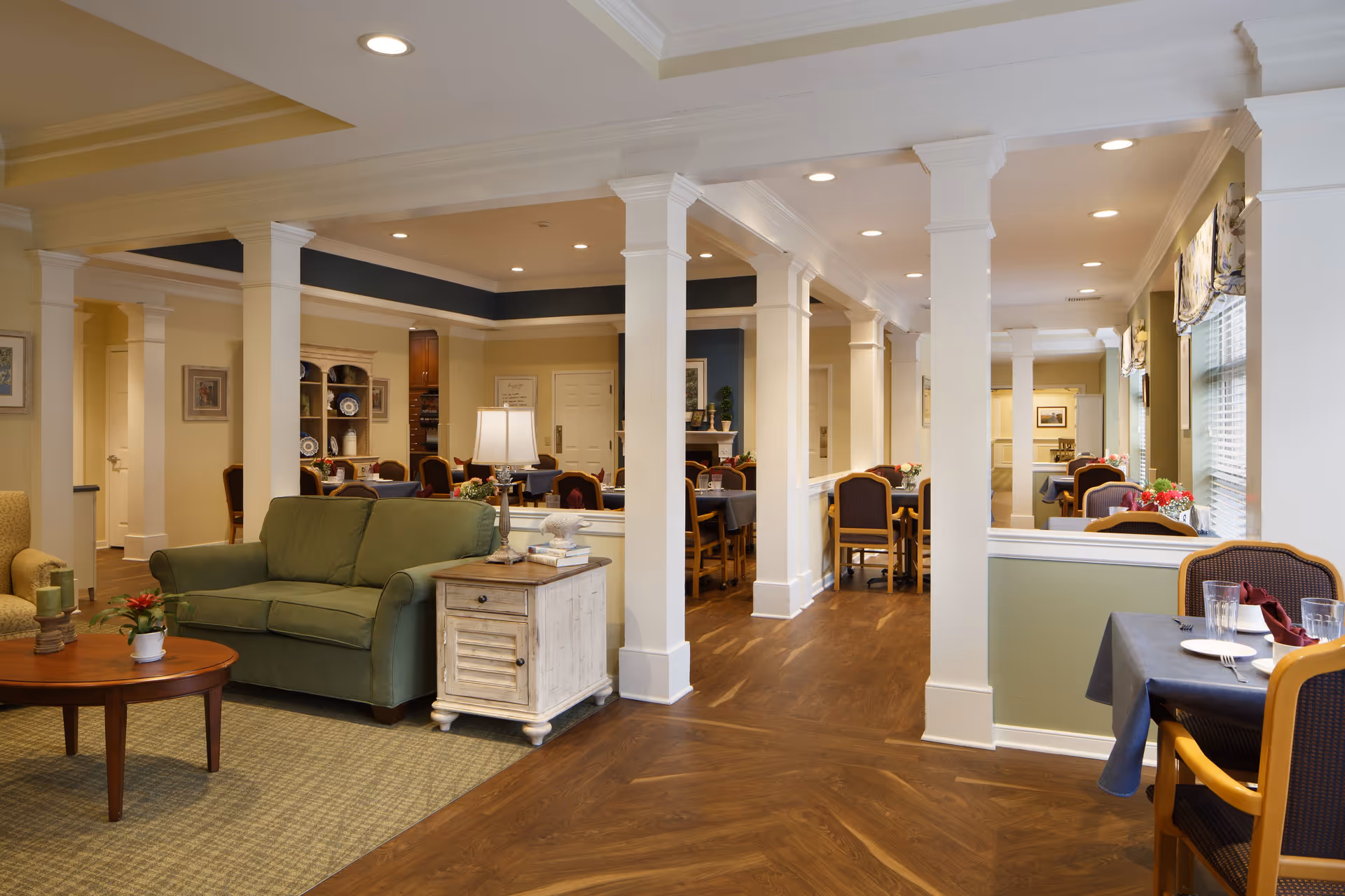 Interior view of a senior living facility showing a cozy seating area with a green couch and wooden side table with a lamp, adjacent to a dining area with multiple tables and chairs set for meals. The space features white columns, warm lighting, and large windows with blinds and valances.