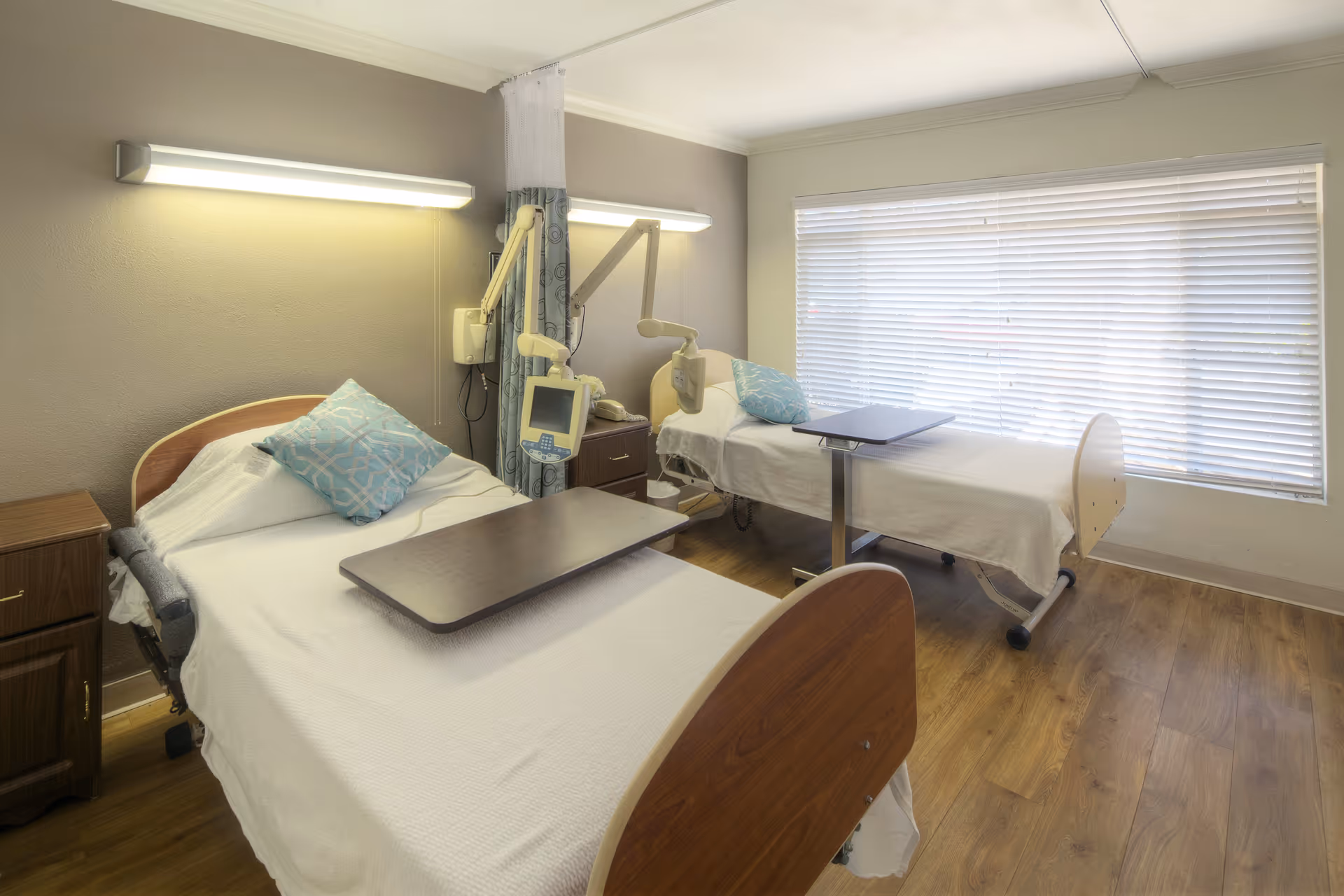A bright room with two hospital-style beds, each with a blue patterned pillow and a movable overbed table. The room has wooden flooring, a large window with blinds, wall-mounted medical equipment, and soft lighting fixtures above each bed.