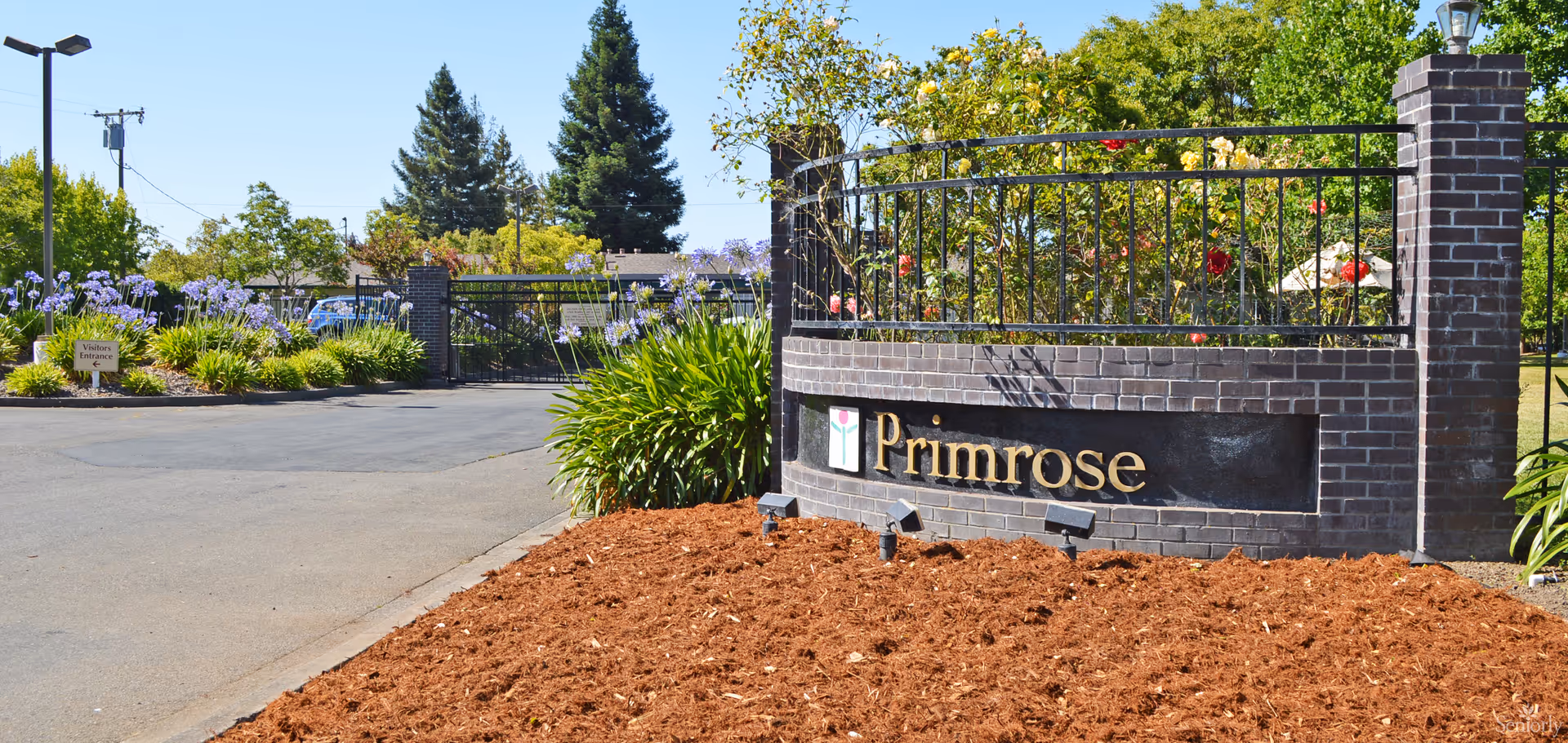 Entrance area of Primrose Alzheimer's Living Inc with a brick sign displaying the name 'Primrose' surrounded by landscaped plants and flowers, a paved driveway, and a gated fence in the background under a clear blue sky.