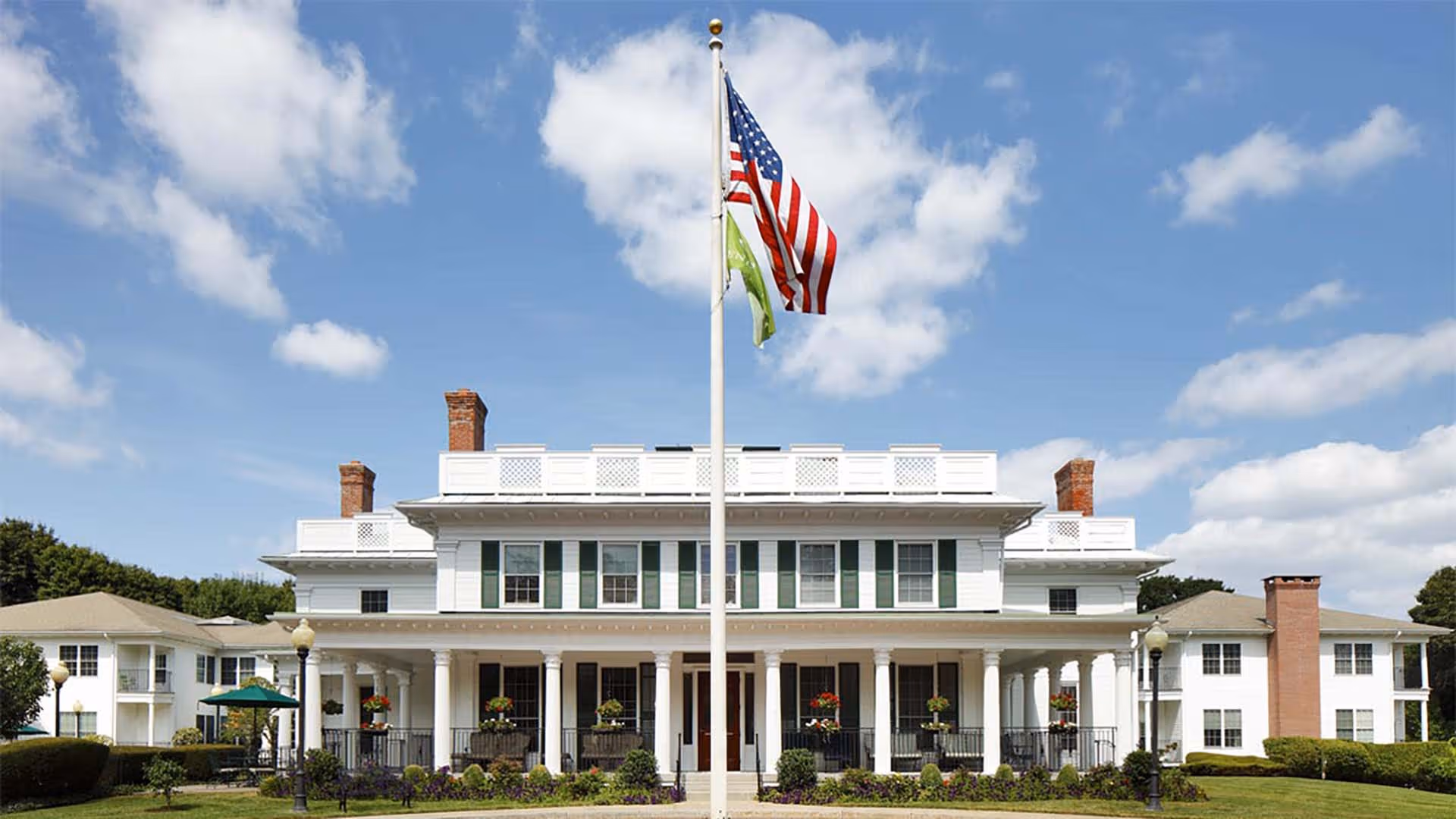 Front exterior view of a large white senior living facility building with green shutters, multiple chimneys, and a wide porch with columns. An American flag and a green flag are flying on a flagpole in front of the building under a partly cloudy blue sky.