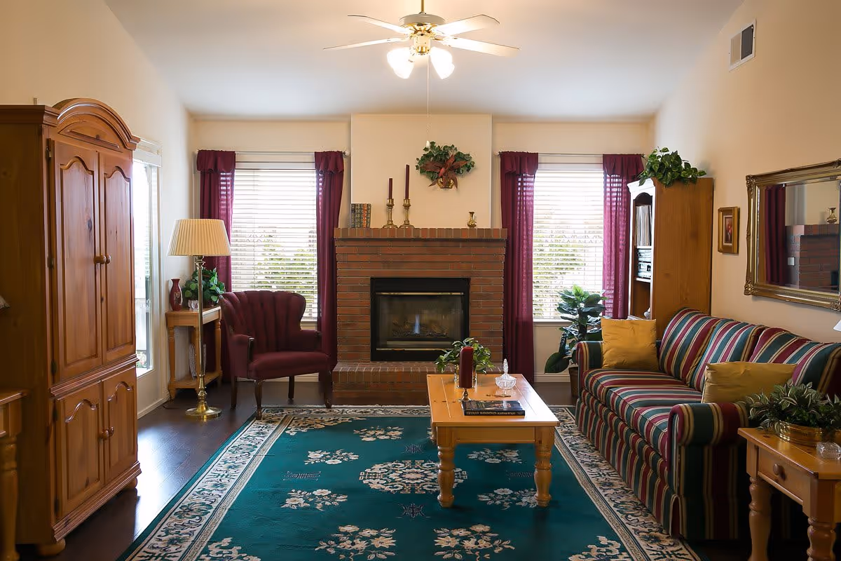 A cozy living room with a striped sofa adorned with yellow pillows, a wooden coffee table with decorative items, a brick fireplace in the center flanked by two windows with burgundy curtains, a burgundy armchair, wooden furniture including a cabinet and side tables, a green patterned rug, and a ceiling fan with lights.