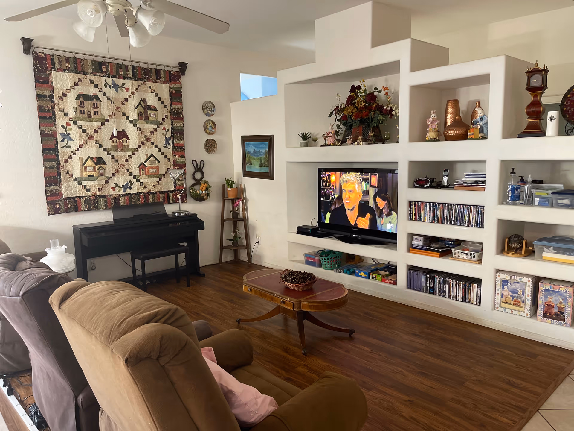 A cozy living room with a brown recliner and a gray sofa facing a built-in entertainment center. The entertainment center has a flat-screen TV displaying a man and a woman, shelves with DVDs, decorative items, and board games. A wooden coffee table with a bowl of pinecones sits on a wood floor. On the left wall, there is a colorful quilt hanging above a black piano with a small bench. A ceiling fan with lights is visible above.