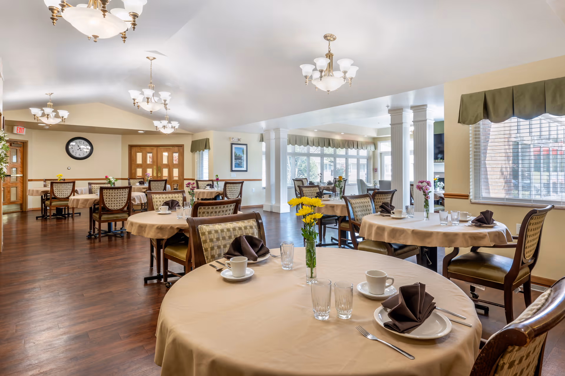 A bright and spacious dining room with round tables covered in beige tablecloths, each set with white plates, cups, glasses, silverware, and folded brown napkins. Yellow and purple flowers in vases decorate the tables. The room has wooden floors, multiple chandeliers, large windows with green valances, and white columns near the windows.