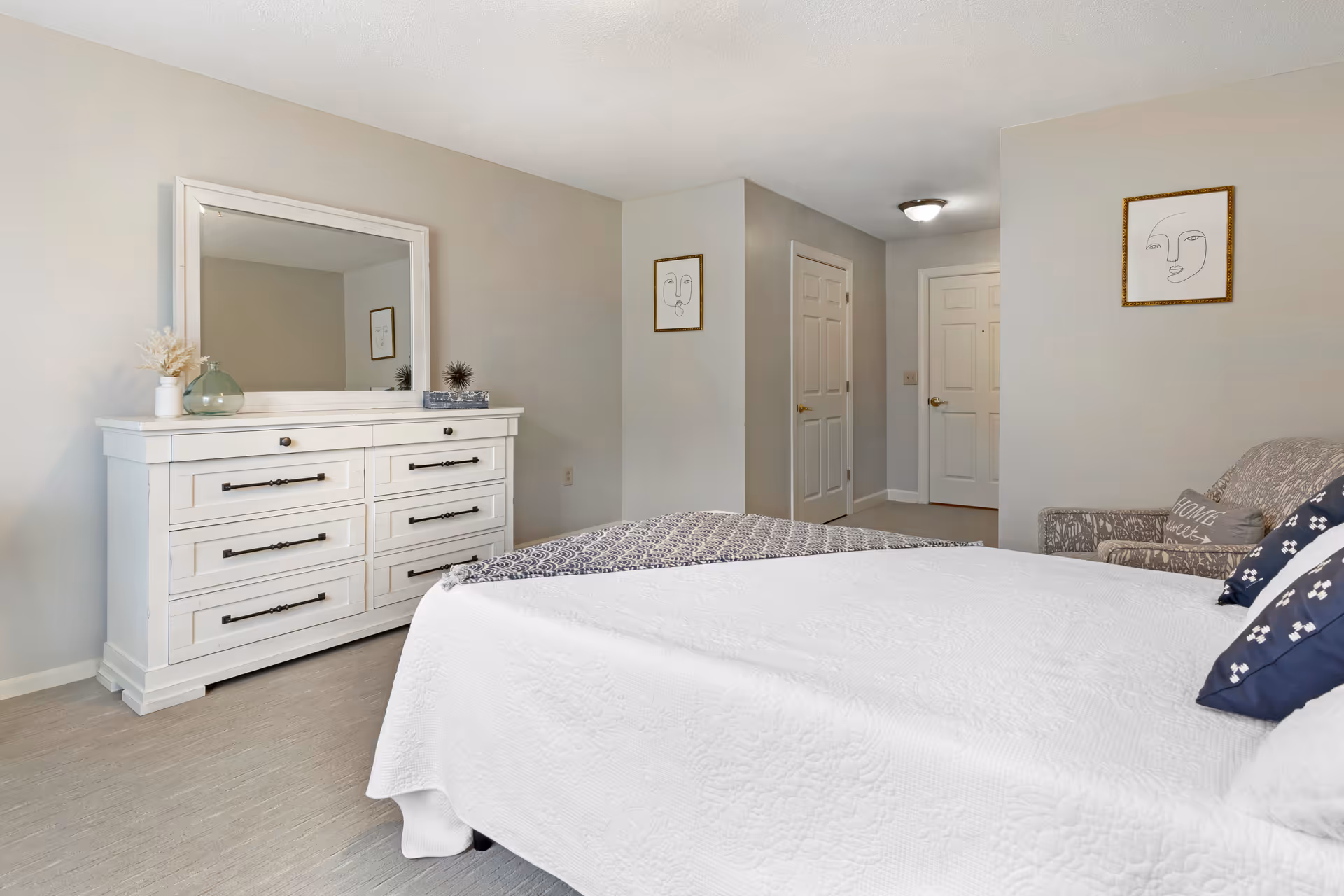 Bright bedroom with a white dresser and mirror, a bed with a white quilt, and a doorway leading to a hallway.