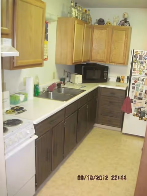 Small galley kitchen with upper and lower wooden cabinets, white countertops, sink, microwave, stove, and a magnet-covered refrigerator.