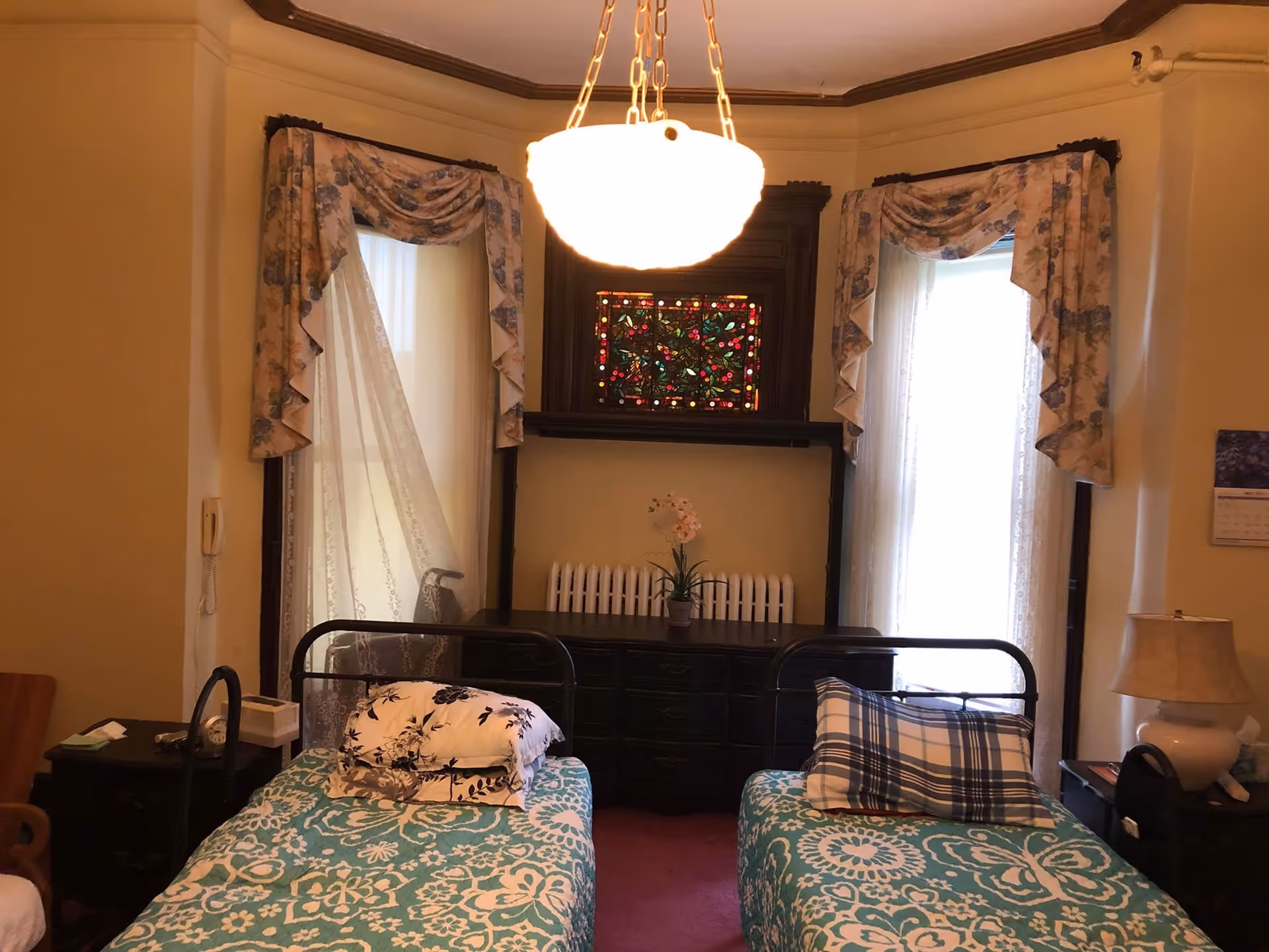 A bedroom with two single beds covered in teal and white patterned bedspreads. Each bed has pillows with different patterns. Between the beds is a dark wooden dresser with a potted plant on top. Behind the dresser is a stained glass window and two tall windows with floral curtains and lace sheers. A hanging light fixture is centered above the beds. There are nightstands on either side of the beds, one with a lamp and the other with a clock and tissue box.