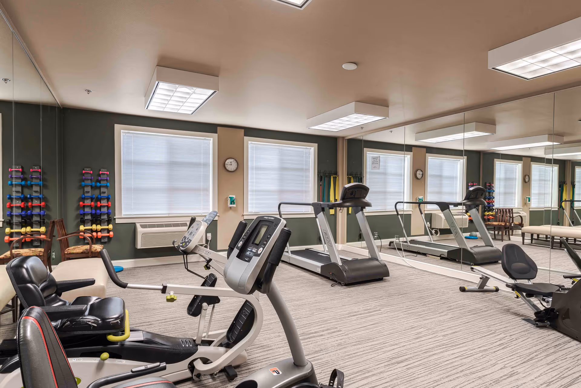 Exercise room with various fitness equipment including a recumbent bike, treadmill, and rowing machine. The room has large windows with blinds, a wall clock, mirrored walls, and racks of colorful dumbbells. There are also chairs and a bench along the walls.