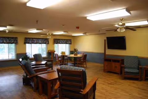 Senior living lounge with wooden chairs, round tables, windows, and a wall-mounted TV.