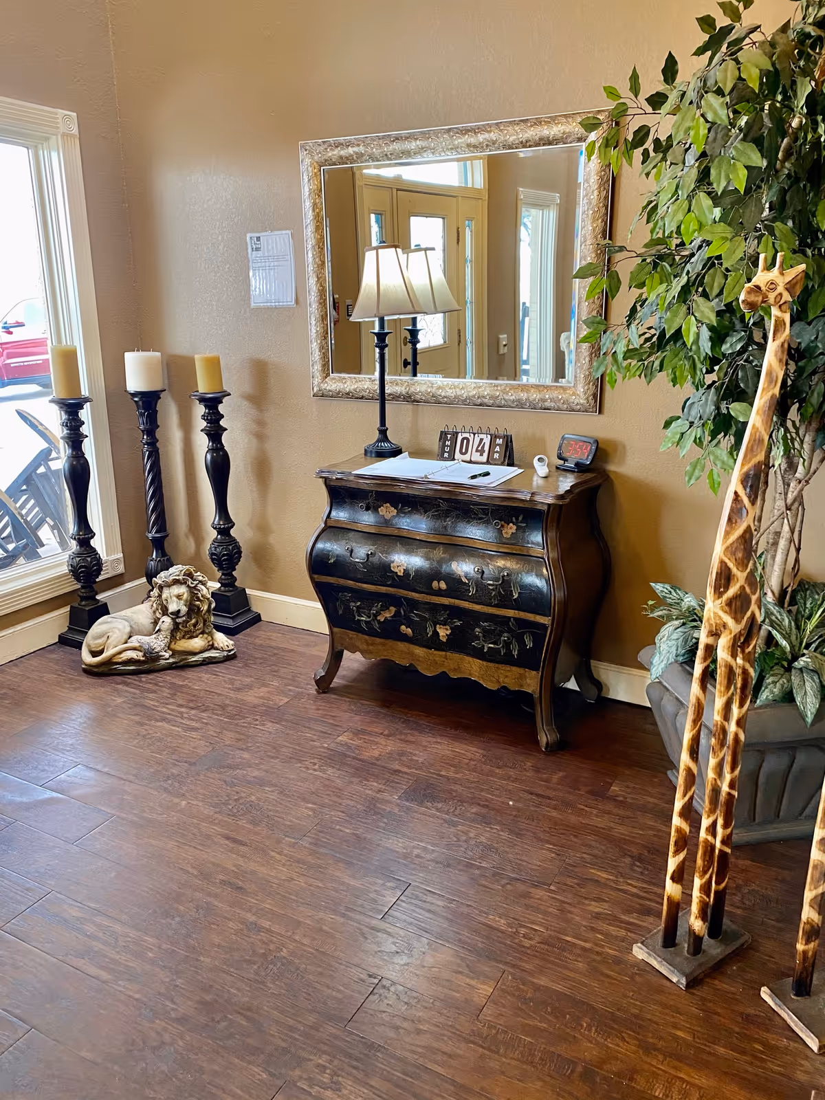 An interior corner of a room with wooden flooring and beige walls. There is a decorative black chest with floral designs, topped with a lamp, a calendar showing March 4, and some papers. Above the chest is a large rectangular mirror with a silver frame. To the left, there are three tall black candle holders with candles and a lion statue lying on the floor. To the right, there is a tall potted plant and two tall wooden giraffe sculptures. A window and a door are reflected in the mirror.