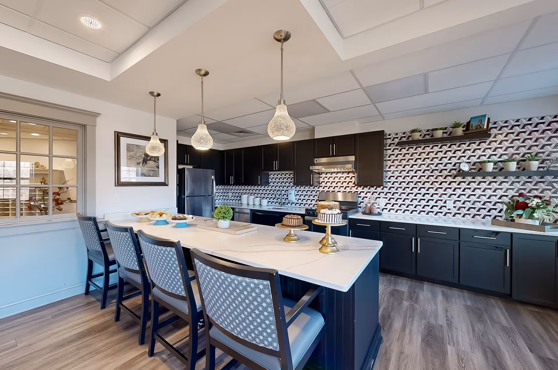 Modern kitchen with a large white island countertop surrounded by four chairs with patterned backs. Three pendant lights hang above the island. The kitchen features dark cabinetry, a stainless steel refrigerator, stove, and a backsplash with a geometric pattern. Shelves on the right wall hold small plants and decorative items.