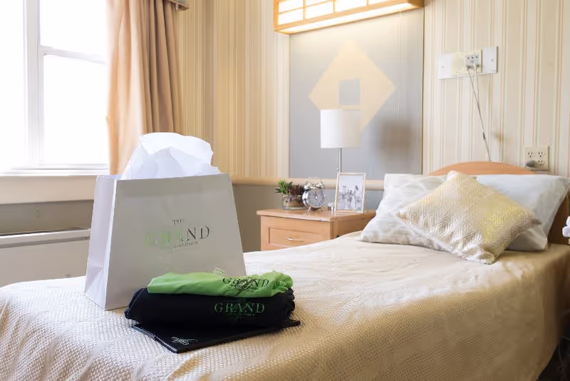A neatly made single bed in a room with beige and white striped wallpaper. On the bed are a white gift bag with tissue paper and folded clothing items with 'The Grand Rehabilitation And Nursing At Queens' logo. Next to the bed is a wooden nightstand with a lamp, a small plant, a clock, and a framed photo. A window with beige curtains is visible on the left side.