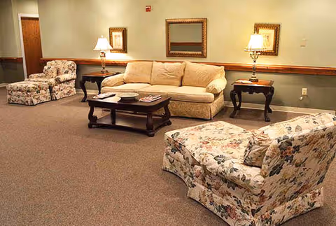 A cozy living room with a beige sofa, two floral-patterned armchairs, a dark wooden coffee table with magazines and plates, two side tables with lamps, and framed artwork on the wall.