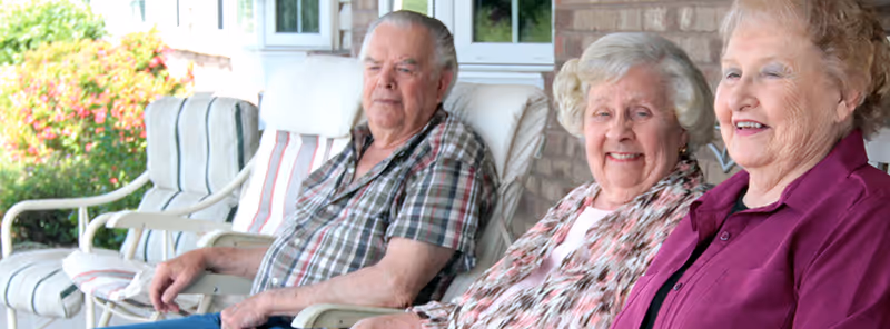 Three elderly people sitting on cushioned chairs on a porch or patio area outside a brick building, smiling and enjoying the outdoor setting with greenery in the background.