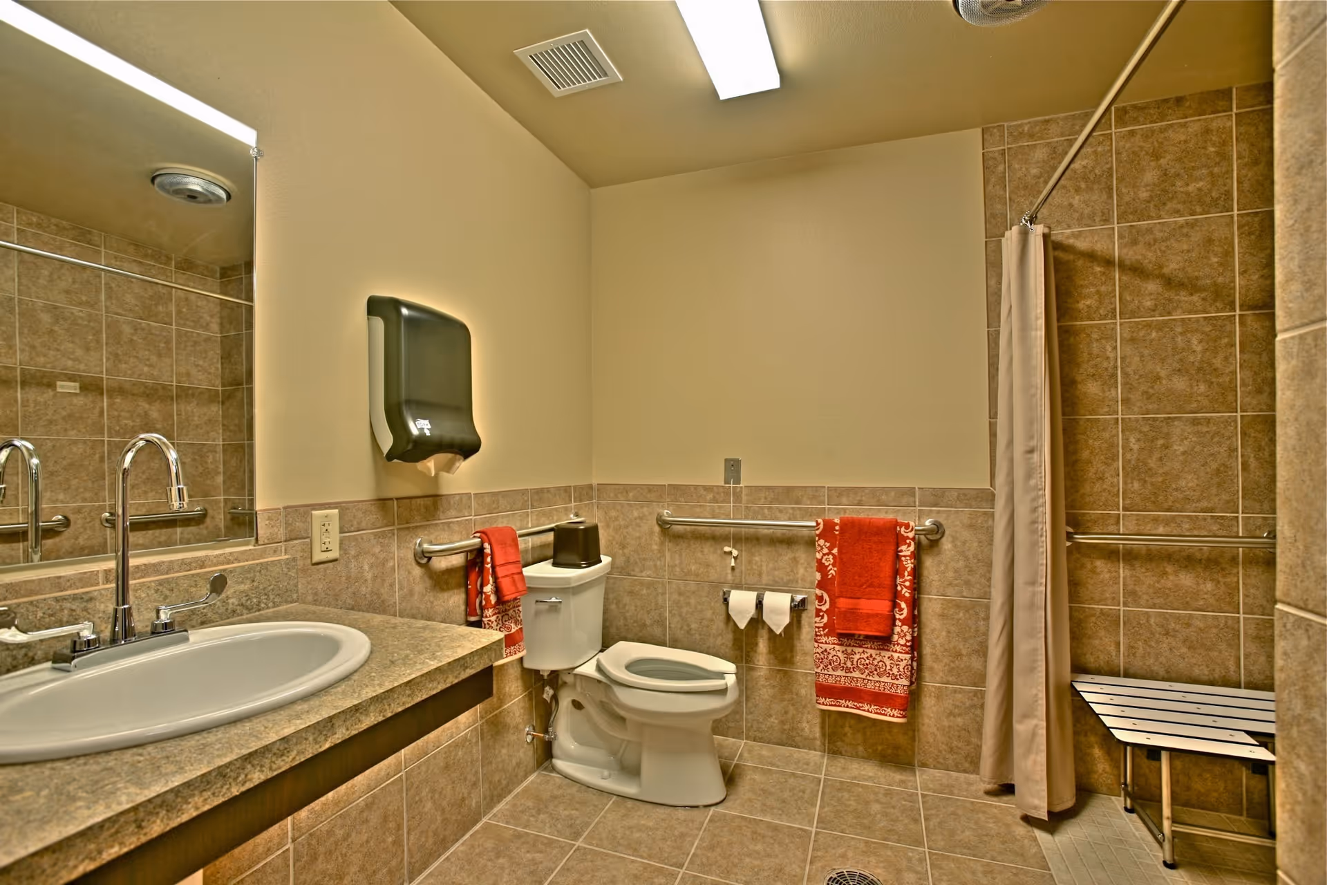 Accessible bathroom with a sink and countertop, toilet with grab bars, shower with bench and curtain, and red towels.