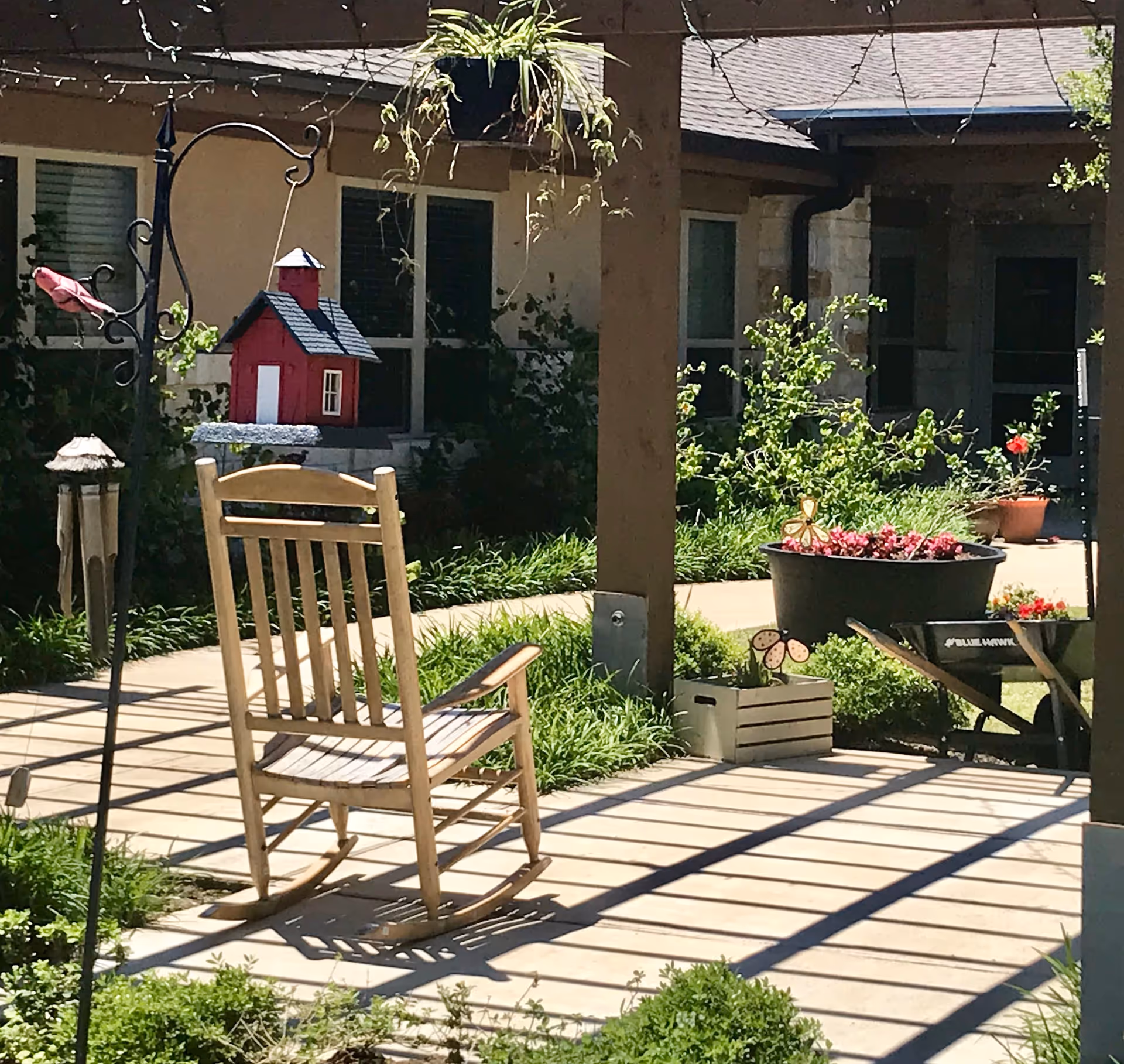 A sunny outdoor patio area with a wooden rocking chair, a hanging red birdhouse, a potted plant with pink flowers, and various garden decorations including a butterfly ornament and a small crate. The background shows windows and part of a building with stone and beige walls.