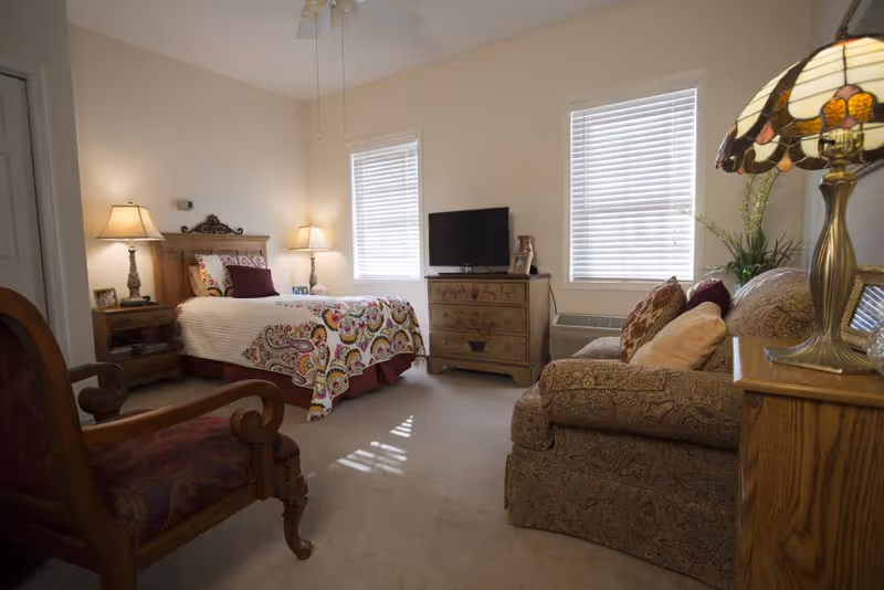 A cozy bedroom with a single bed covered in a colorful patterned quilt, two bedside tables with lamps, a wooden dresser with a TV on top, a patterned armchair, and a patterned sofa with cushions. Two windows with white blinds allow natural light into the room.