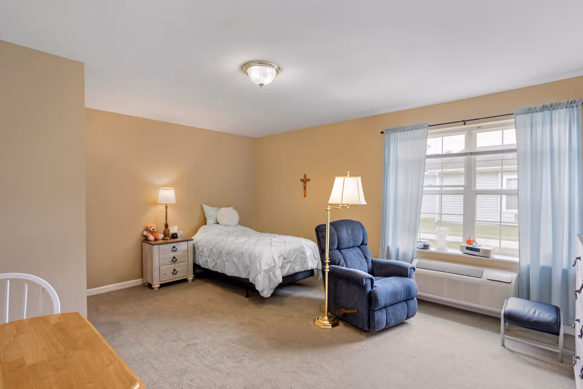 A senior living facility bedroom with beige walls and carpet. The room features a single bed with white bedding, a wooden nightstand with a lamp and small items, a blue recliner chair with a floor lamp beside it, a window with light blue curtains, and a small stool near the window. There is a wooden table and chair partially visible in the foreground.