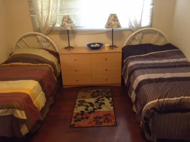 A bedroom with two single beds on either side of a wooden dresser. Each bed has a striped comforter, one in shades of brown and yellow, the other in shades of brown and gray. On top of the dresser are two lamps with patterned shades and a blue bowl with decorative items. A small floral rug is placed on the wooden floor between the beds. Light filters in through a window with sheer curtains behind the dresser.