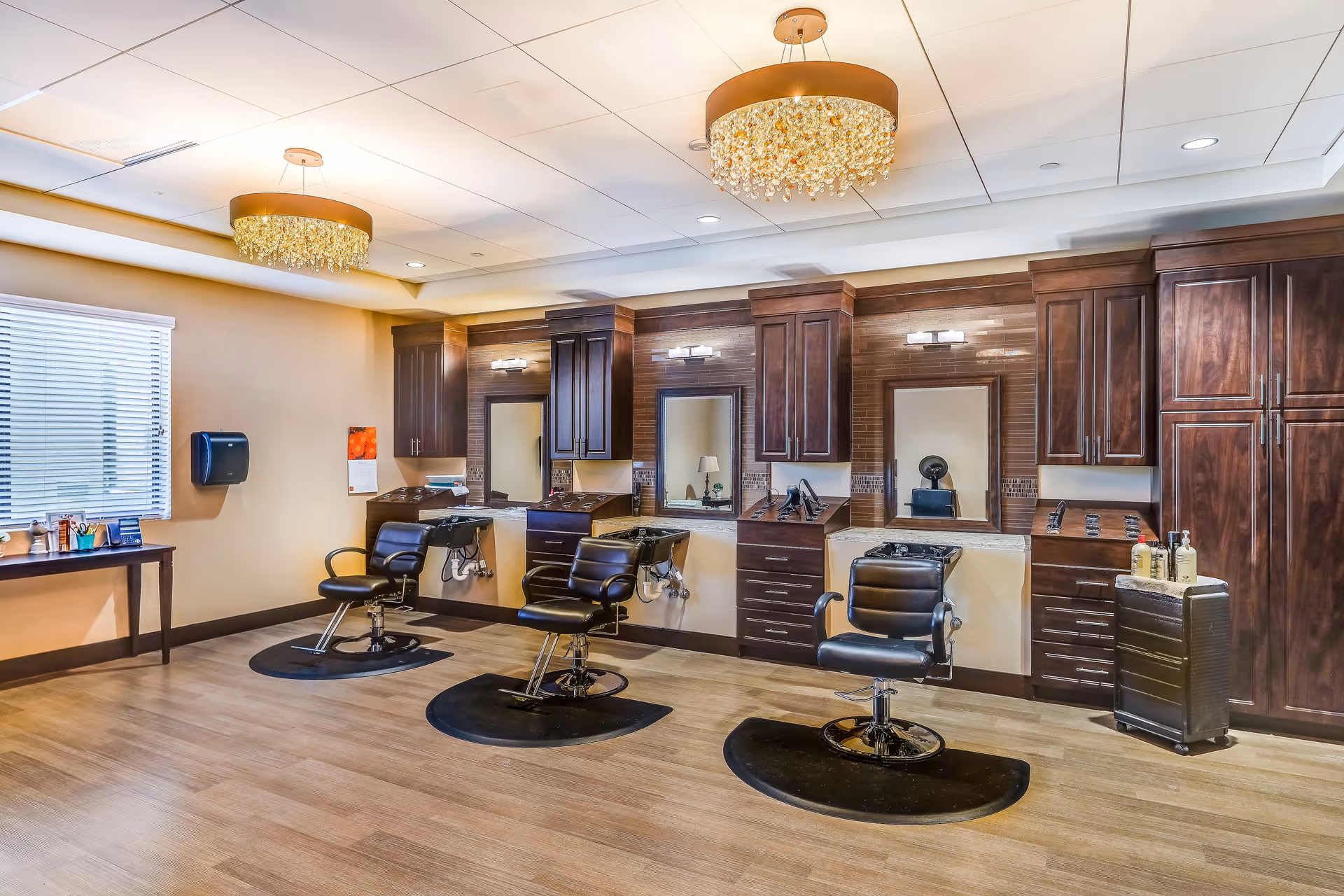 Interior view of a salon area with three black salon chairs in front of mirrors and wooden cabinetry. The room has light wood flooring, two decorative ceiling lights, and a window with blinds on the left side.