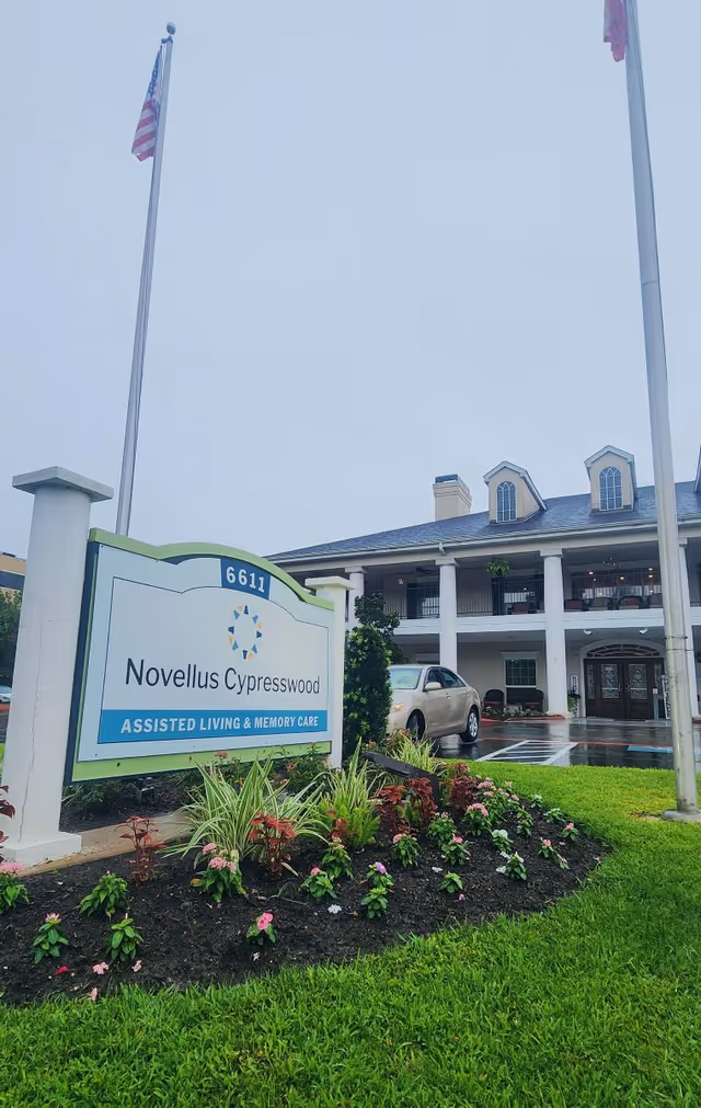 Front entrance of Novellus Cypresswood assisted living facility with its sign, landscaped flowerbed, flagpoles, and a two-story building.