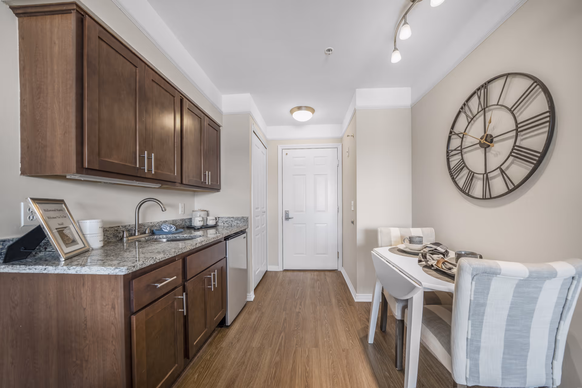 Small kitchenette with wooden cabinets, granite countertop, sink and a two-seat dining table under a large wall clock.