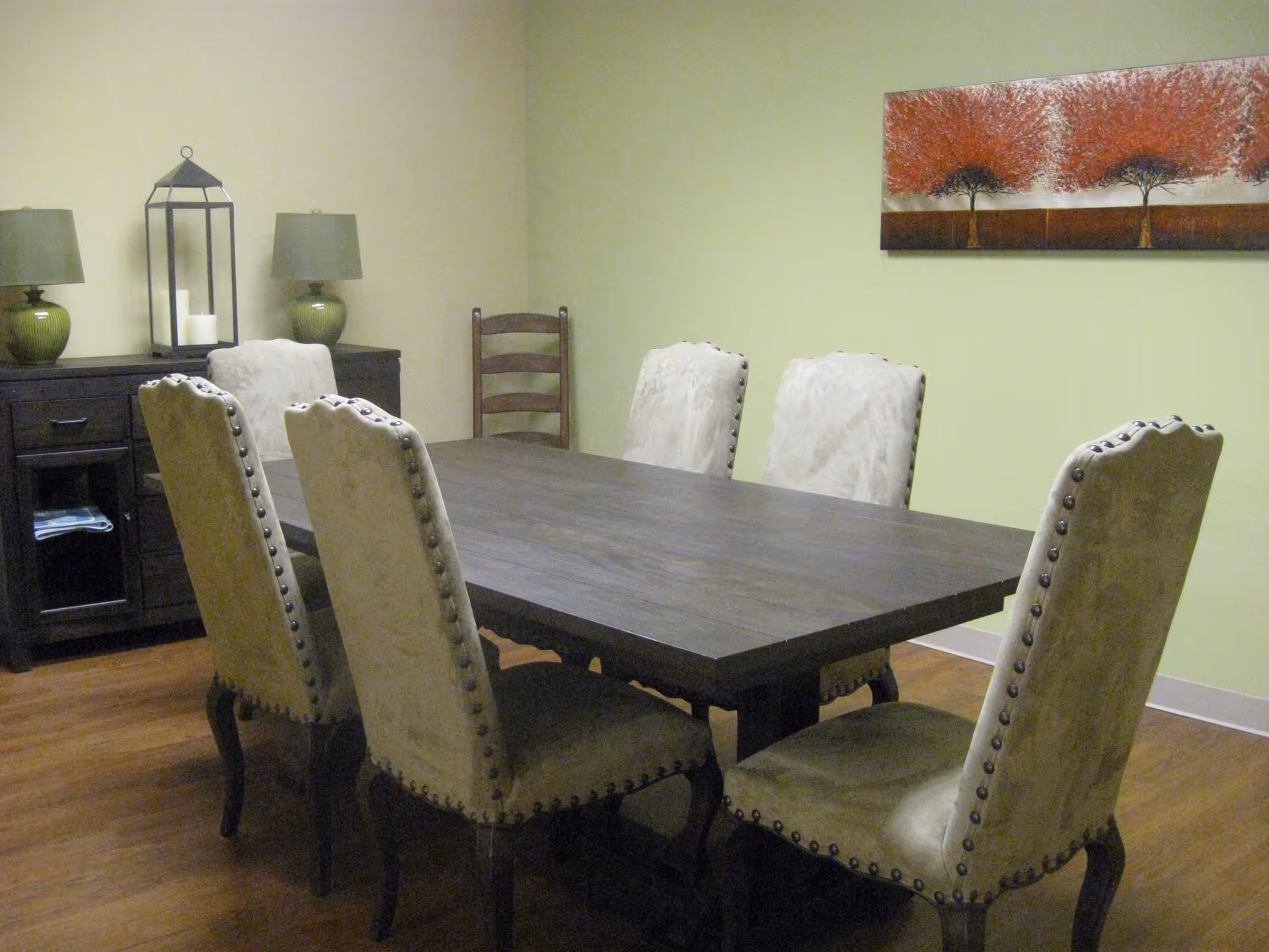 A dining room with a dark wooden table and six upholstered nailhead chairs, a sideboard with lamps, and a tree painting on a pale green wall.