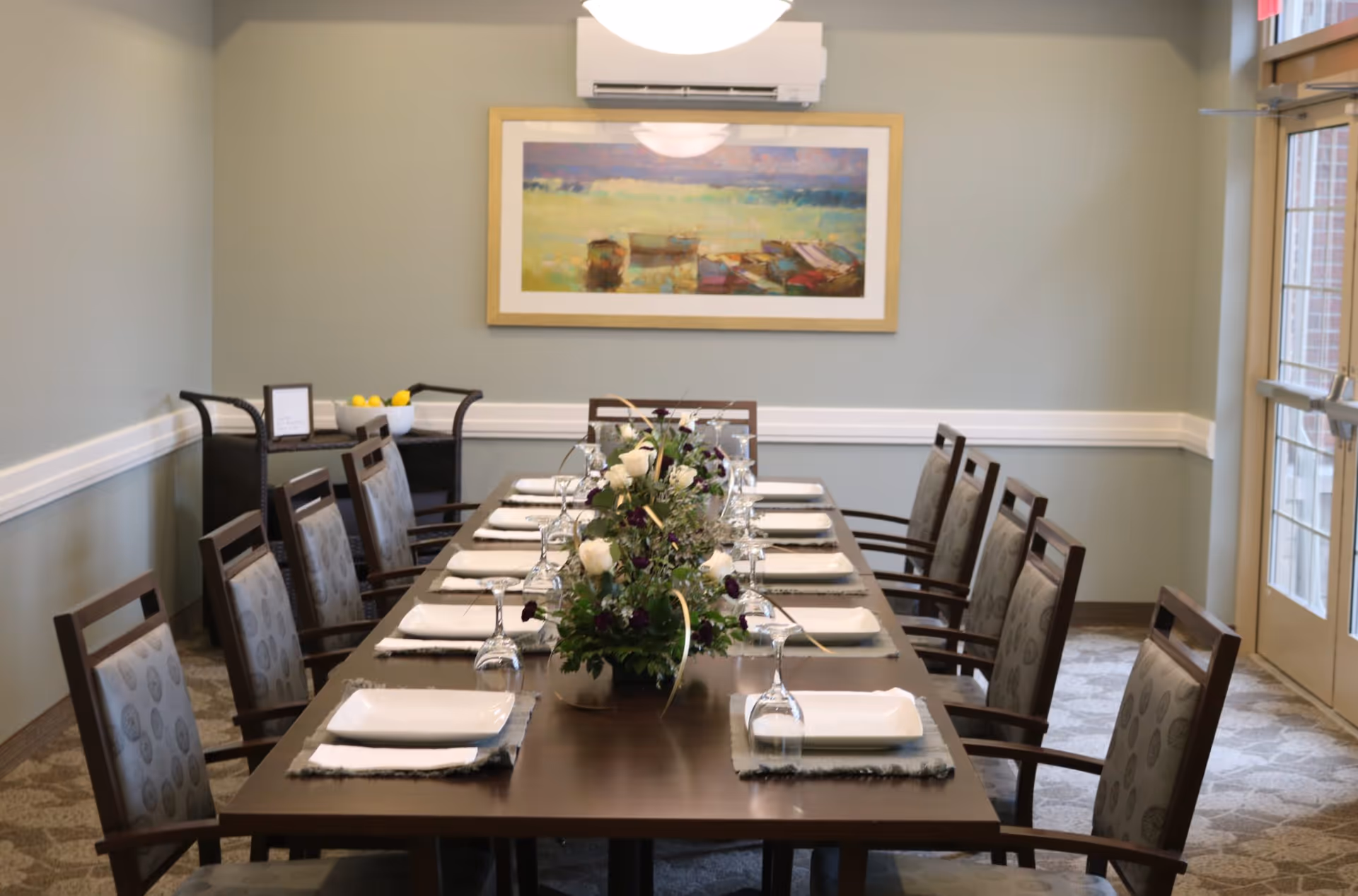 A dining room with a long wooden table set for ten people. Each place setting includes a white plate, a napkin, and an upside-down wine glass. A floral centerpiece with white and dark purple flowers is placed in the middle of the table. The room has light green walls, a framed painting of boats on the water, a wall-mounted air conditioning unit, and a windowed door letting in natural light.