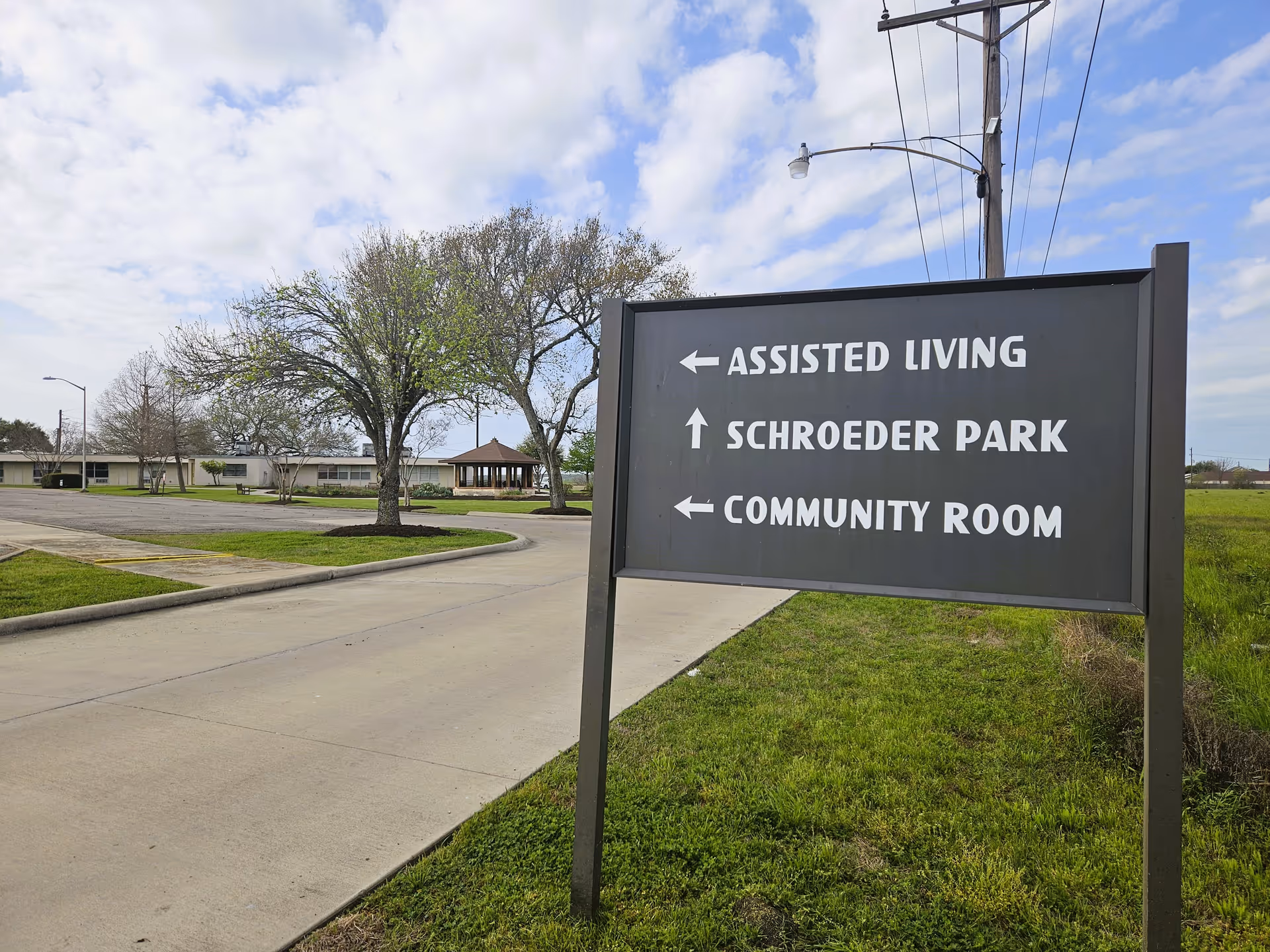 Directional sign on the grounds pointing to Assisted Living, Schroeder Park, and Community Room with the facility building and gazebo in the background.