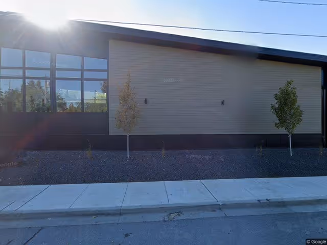 Exterior side view of a building with beige siding and black trim, two small trees planted in a gravel bed along the sidewalk, and sunlight shining from the upper left corner.