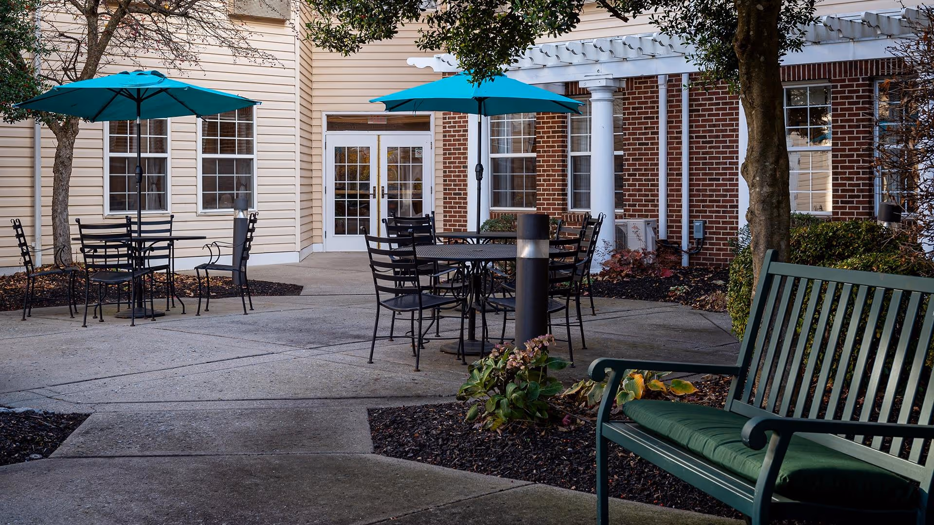 Outdoor patio area at a senior living facility with several black metal tables and chairs, each shaded by teal umbrellas. There is a green bench with a cushion in the foreground, surrounded by landscaped garden beds and trees. The building exterior features beige siding and red brick with white columns and multiple windows.